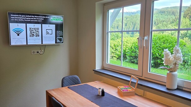 Una stanza con un tavolo di legno, due sedie, una finestra con vista sul verde e uno schermo a parete che mostra codici QR.