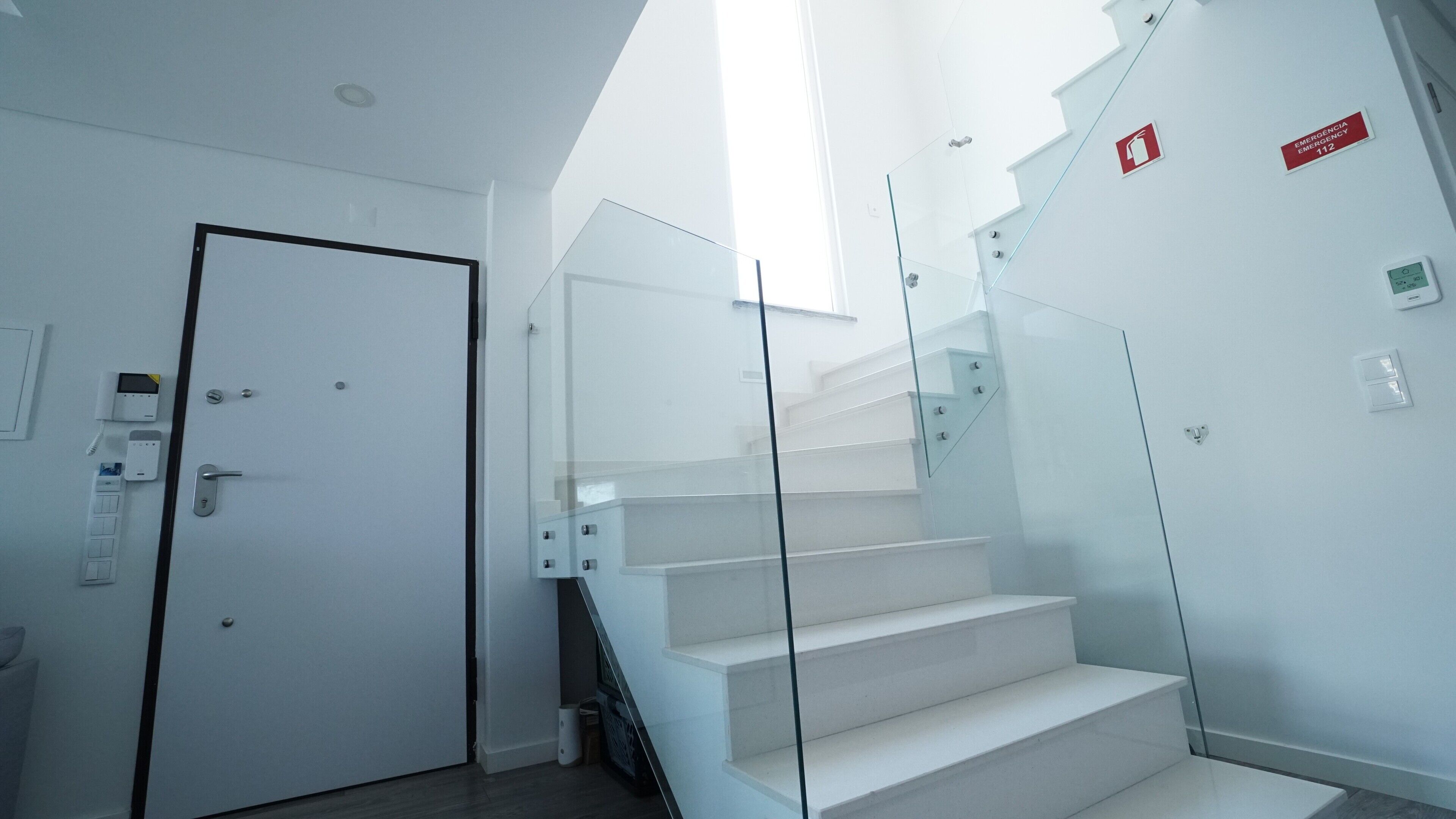 Un interior moderno con una escalera de cristal, una puerta blanca y señalización de emergencia.