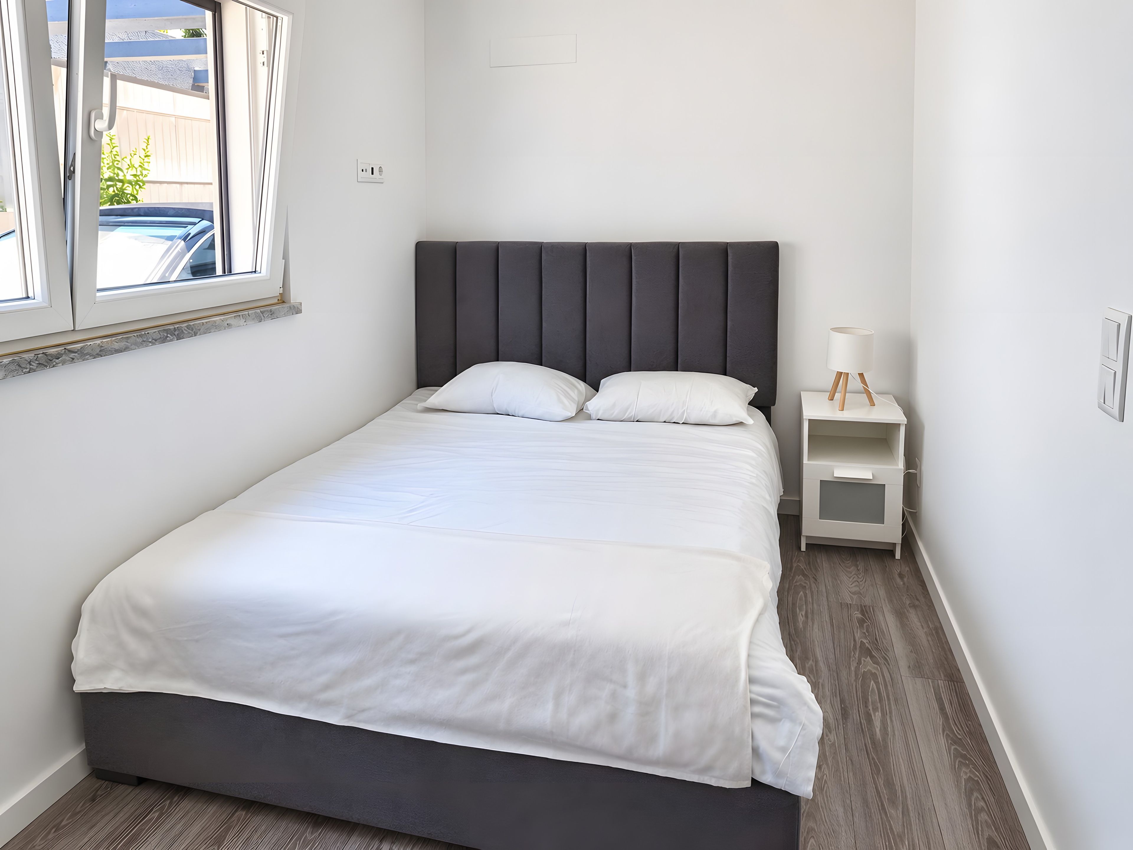 Una cama bien tendida con un cabecero oscuro en una habitación con paredes blancas y una mesita de noche pequeña con una lámpara.