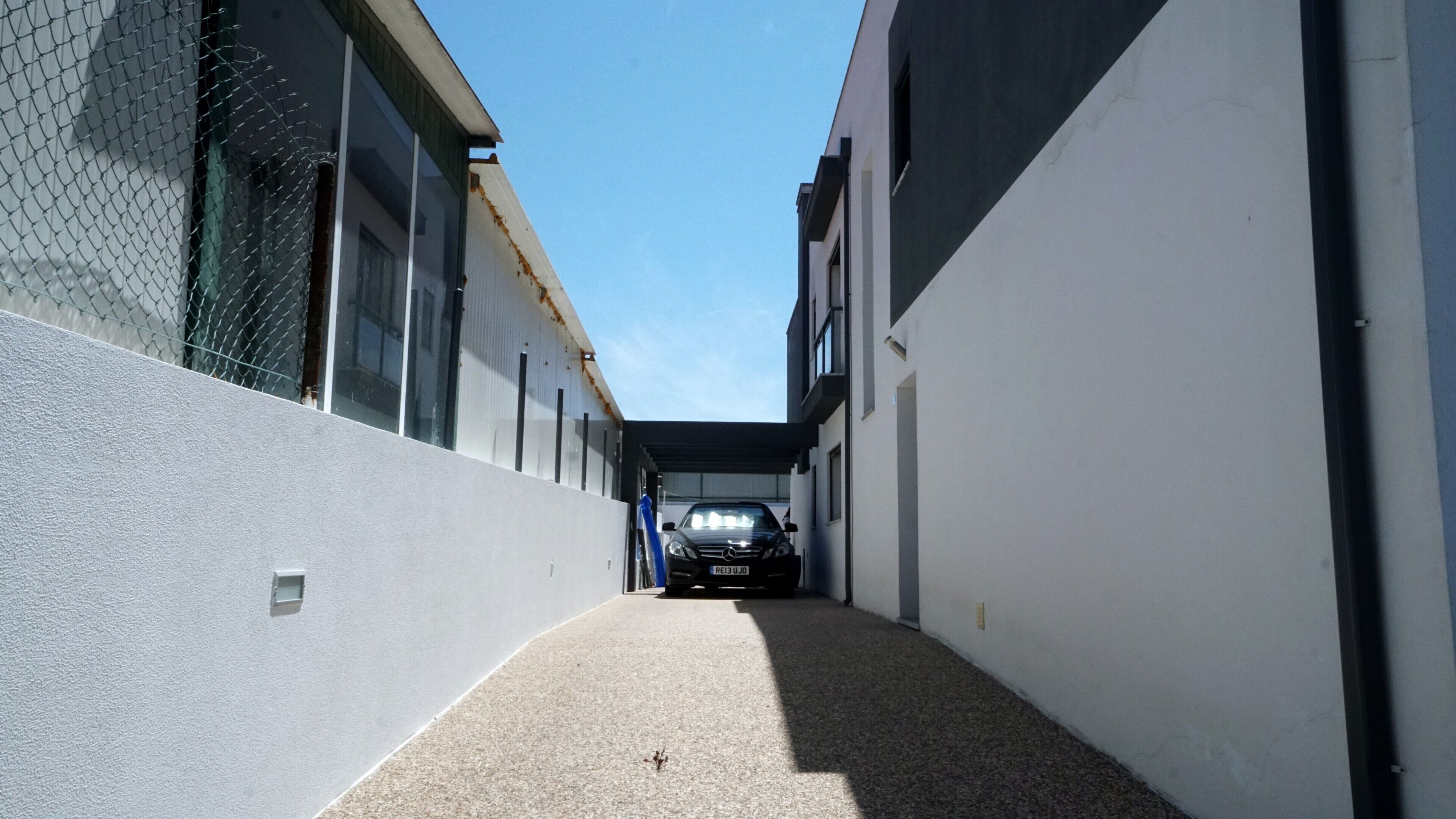 Un estrecho callejón con un coche al final, flanqueado por edificios con grandes ventanales y una cerca de malla metálica en un lado.