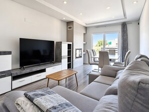 A modern living room with a flat-screen TV, a sofa, a coffee table, and a dining area with chairs.