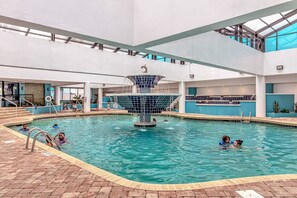An indoor swimming pool with a central fountain, surrounded by a tiled deck and a high ceiling with skylights. - Landmark 907 Ocean view studio (Myrtle Beach)