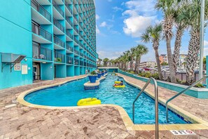 A pool area with a blue building, palm trees, and a clear sky. - Landmark 907 Ocean view studio (Myrtle Beach)