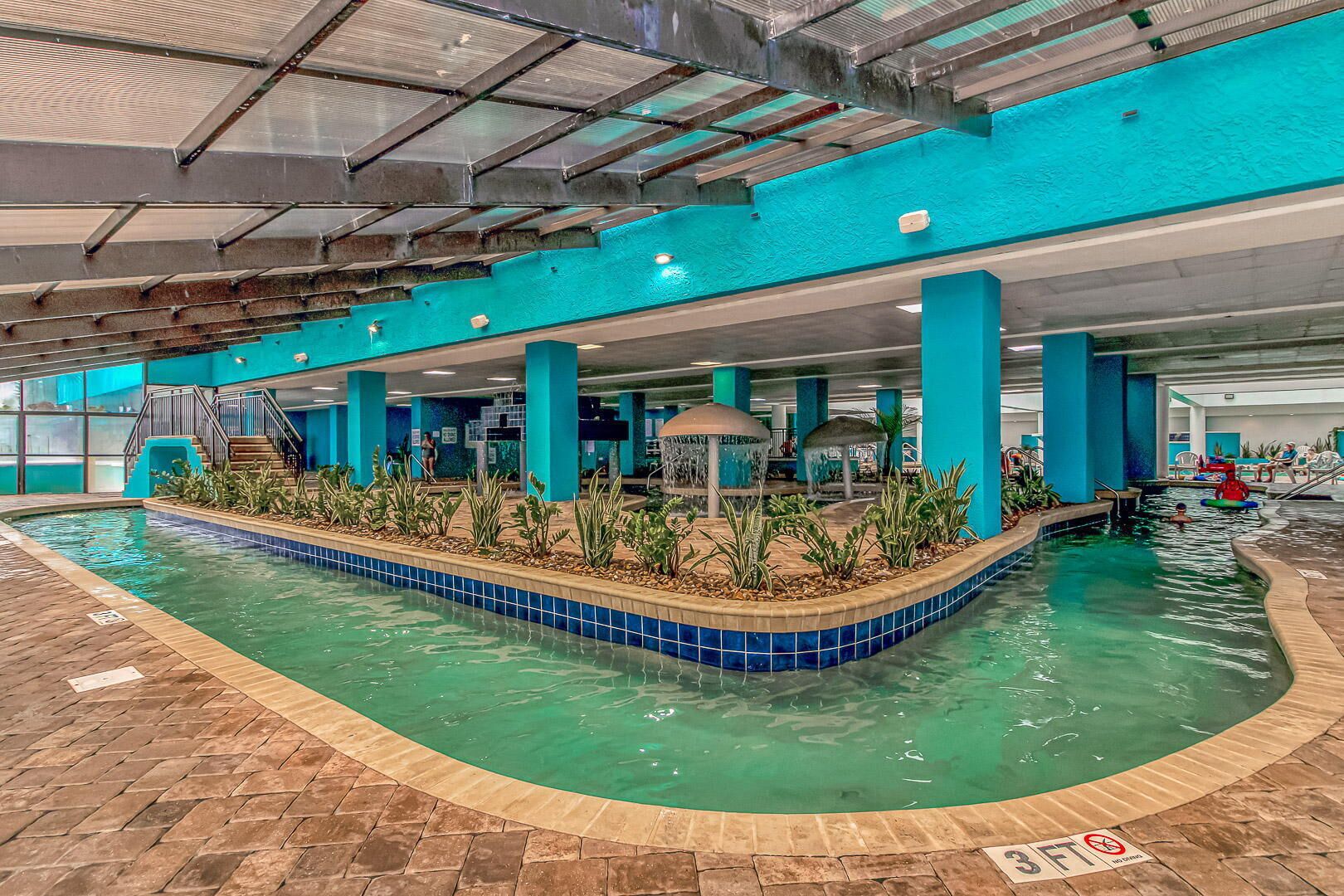 An indoor swimming pool with a tiled edge, a central island with plants, and a covered seating area.