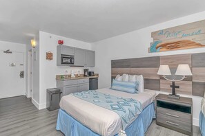 A hotel room with a bed, bedside table, lamp, and a kitchenette area. - Landmark 907 Ocean view studio (Myrtle Beach)