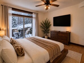 A bedroom with a large bed, a ceiling fan, a TV mounted on the wall, a wooden dresser, and a window with a view of a snowy landscape.
