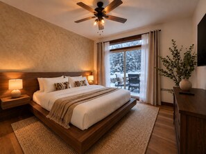 A bedroom with a large bed, a ceiling fan, a nightstand with a lamp, and a view of a snowy outdoor area through a sliding glass door. - Tremblant Prestige - Les Eaux 217-5A (Mont-Tremblant)