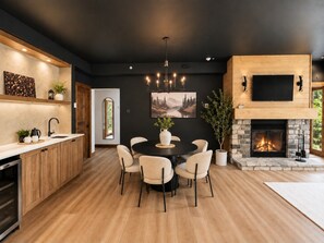 A modern kitchen with a central island, a dining area with a round table and chairs, a fireplace, and a large flat-screen TV mounted on a wooden wall. - Tremblant Prestige - Les Eaux 217-5A (Mont-Tremblant)