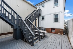 A wooden deck with a metal staircase, a picnic table, and a white building with windows. - Half Block to Beach | King Bed (Seaside Heights)