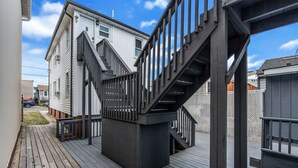 Um deck residencial com piso de madeira, escada de metal com corrimão e uma casa branca ao fundo.
