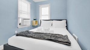 Um quarto com uma cama, duas janelas com persianas, uma mesa de cabeceira e um aquecedor fixado na parede.