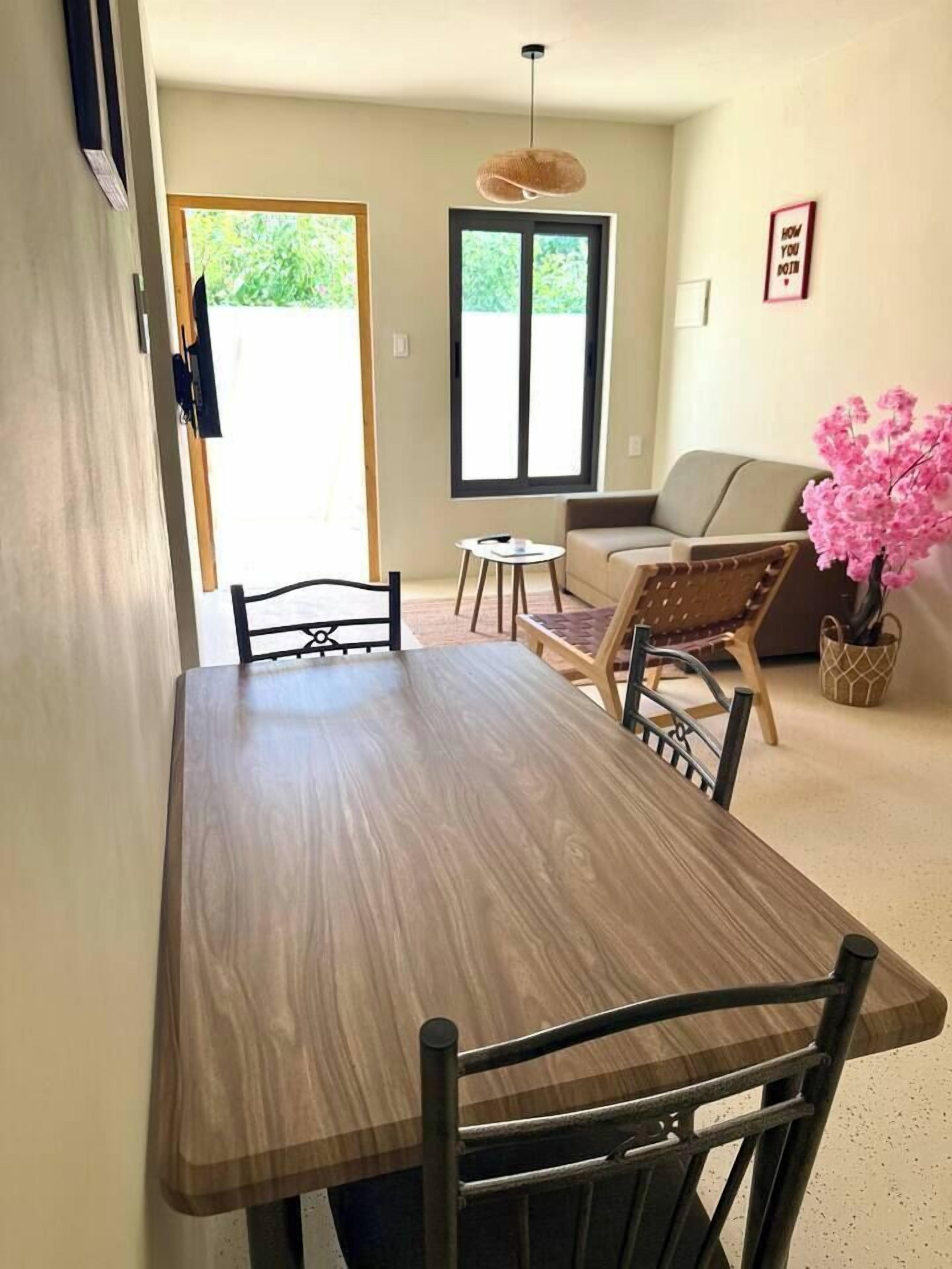 Un comedor con una mesa de madera, sillas negras, una mesita redonda y un sofá. Se observa una ventana con vistas a zonas verdes.