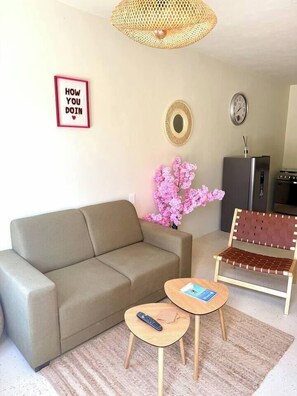 Uma sala de estar com sofá, uma pequena mesa de madeira, uma cadeira de fibra natural, um relógio e um espelho decorativo.