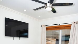 Ein Schlafzimmer mit einem Bett, einem Deckenventilator, einem an der Wand montierten Fernseher und einem Fenster mit Vorhängen.