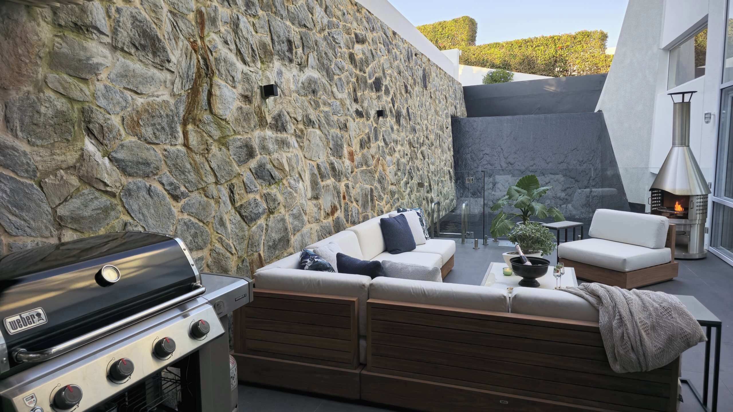 Un patio al aire libre con pared de piedra, una parrilla Weber, un sofá y una silla de lounge.