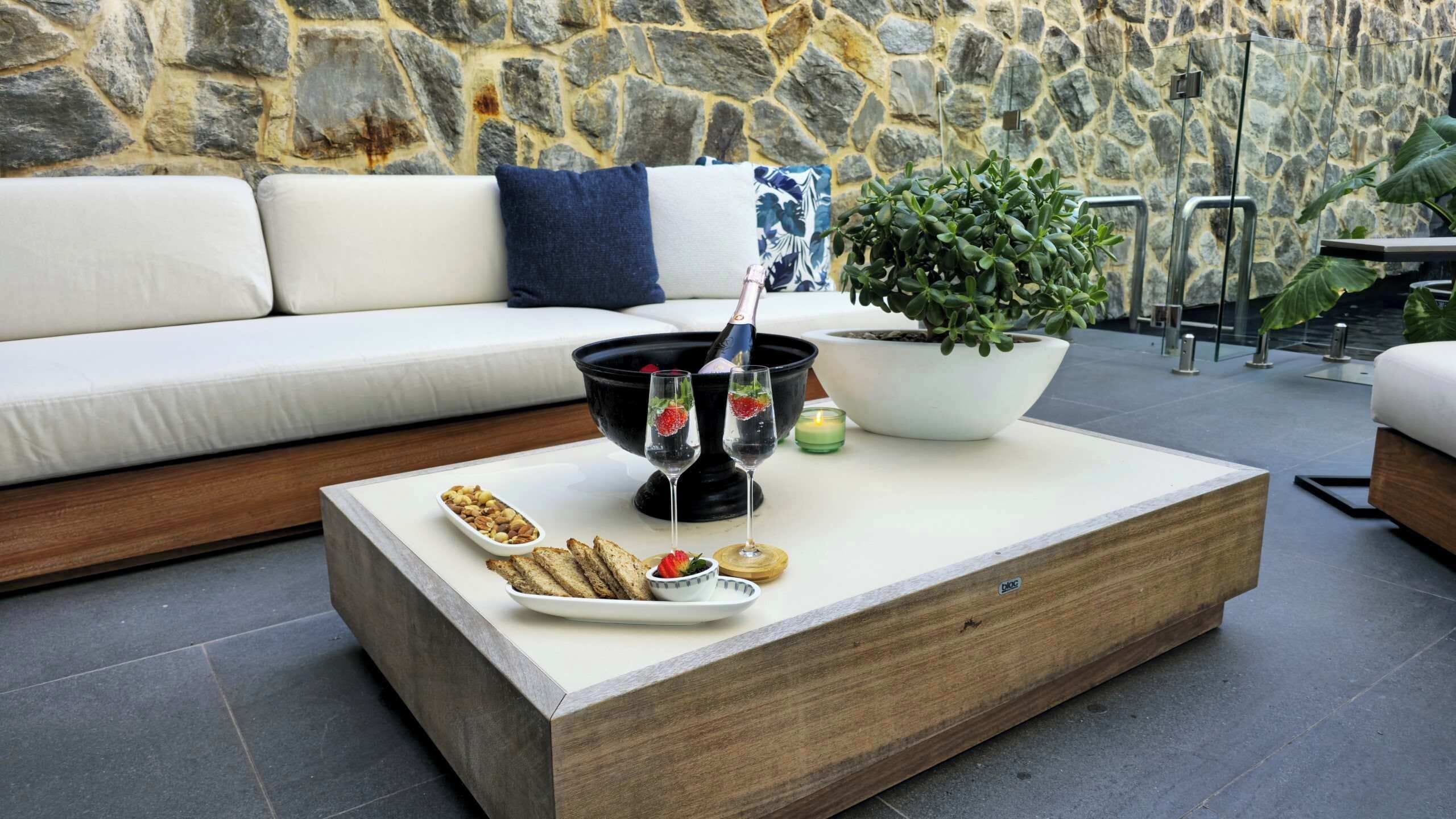 Uma área de estar moderna ao ar livre, com um muro de pedra ao fundo, uma mesa de centro de madeira com petiscos, uma planta em vaso e uma taça de champanhe.