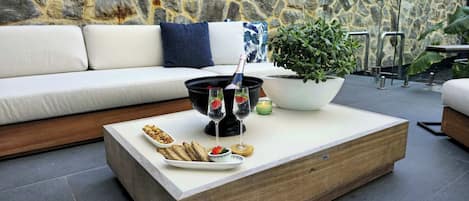 A modern outdoor seating area with a stone wall backdrop, a wooden coffee table with snacks, a potted plant, and a glass of champagne.