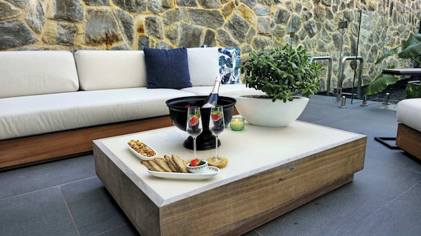 A modern outdoor seating area with a stone wall backdrop, a wooden coffee table with snacks, a potted plant, and a glass of champagne.