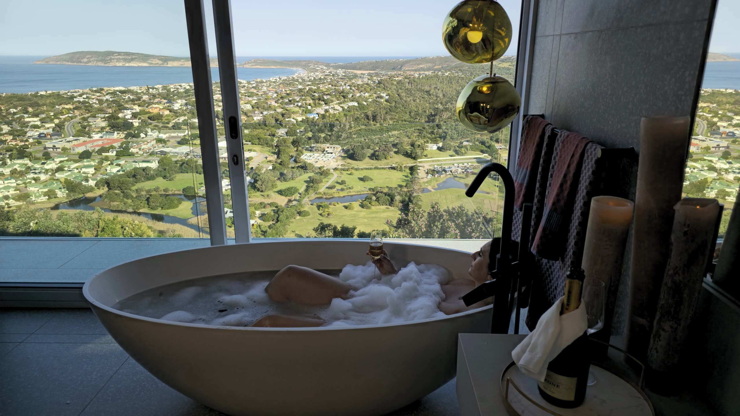Une grande baignoire remplie de mousse, une vue sur un paysage côtier et une bouteille de vin posée sur une table.