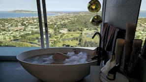 A large bathtub filled with bubbles, a view of a coastal landscape, and a bottle of wine on a table.