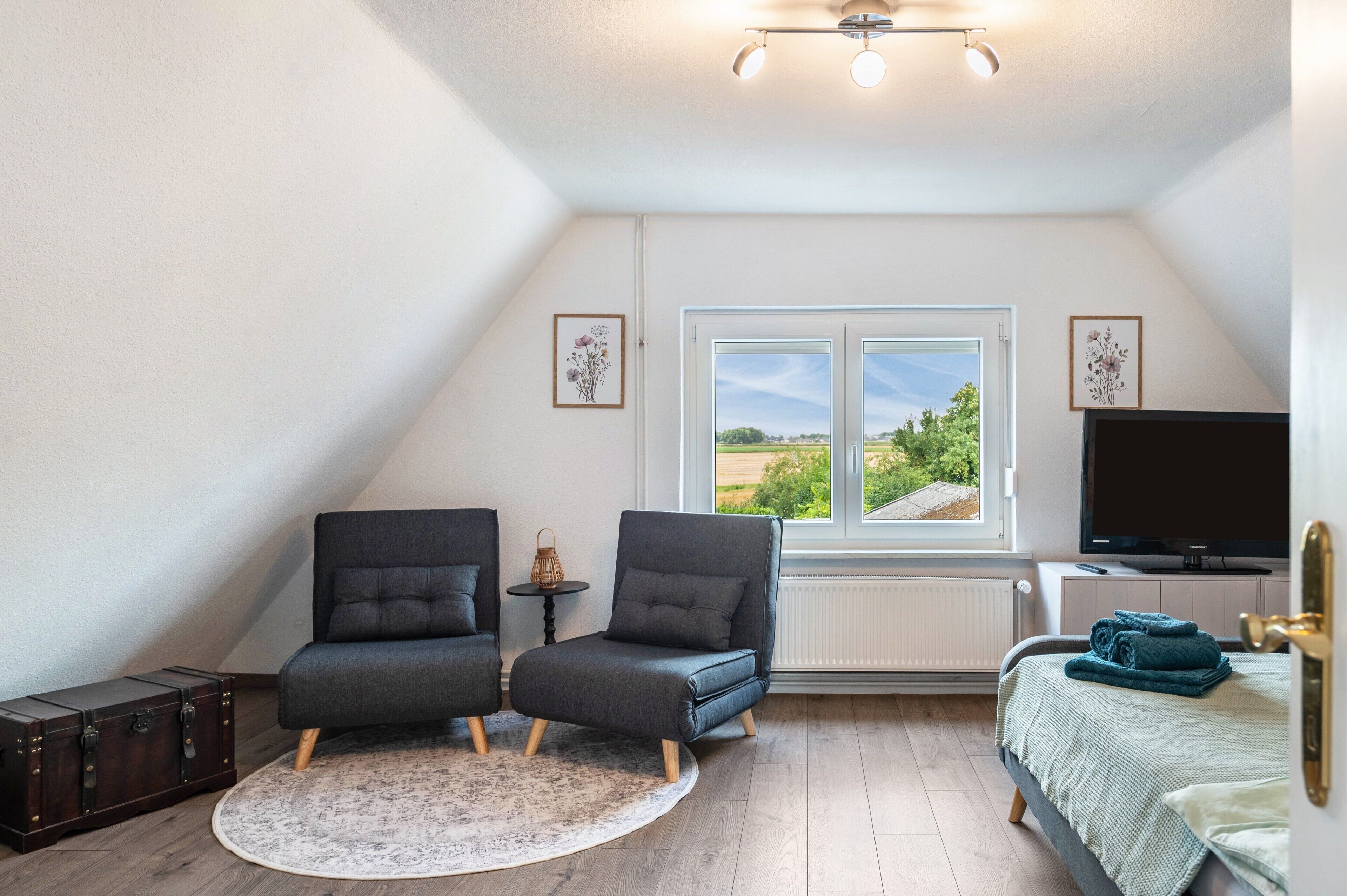 A bedroom with two grey armchairs, a small round table, a bed with a green blanket, a TV, and a window with a view of greenery.