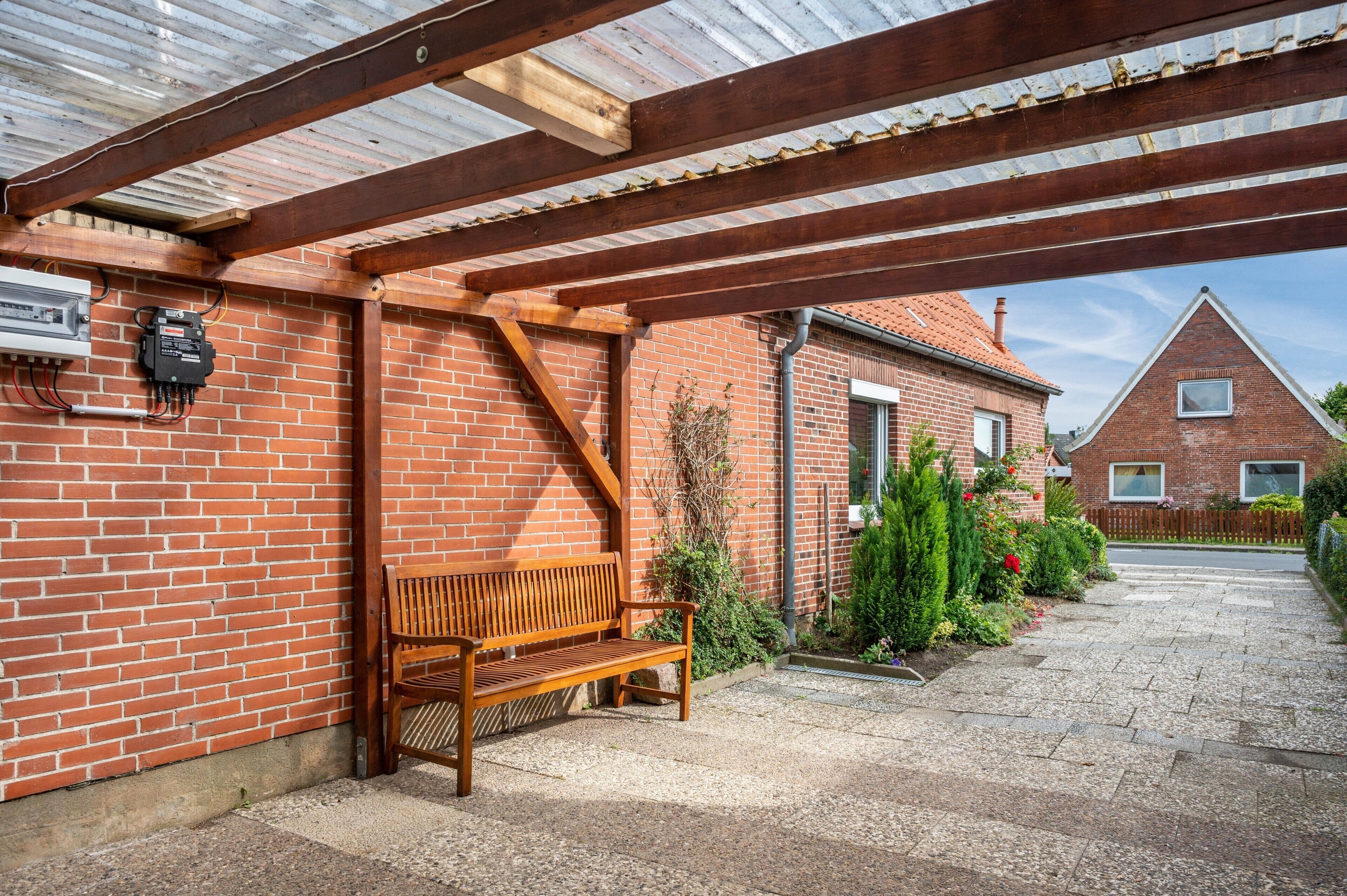 A covered outdoor area with a wooden bench, brick walls, and a gravel driveway leading to a house with a pitched roof.