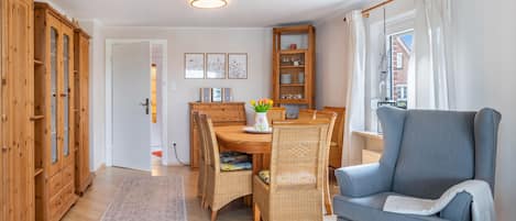 A dining area with a wooden table and chairs, a blue armchair, a wooden cabinet, and a window with curtains.
