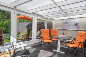 Un solárium con techo de vidrio, mobiliario exterior con sillas naranjas, una mesa y un cuadro en la pared.