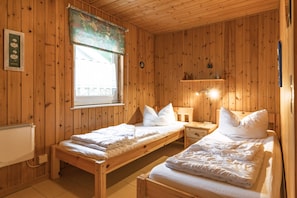 Ett rum med träpanel på väggarna, två enkelsängar, ett element, ett fönster med gardin och en liten hylla med diverse föremål.