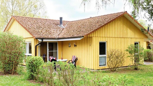 A yellow building with a tiled roof, a front porch with two chairs, and a small table. - Doppelbungalow im Skandinavischen Stil mit Terrasse im Ferienpark Mirow (Mirow)