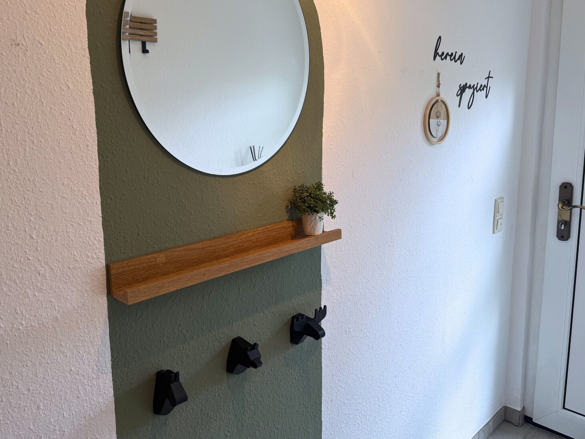 Ein Zimmer mit einem runden Spiegel, einem Holzboden mit einer Pflanze und einer Tür mit einem Wandleuchtschalter.