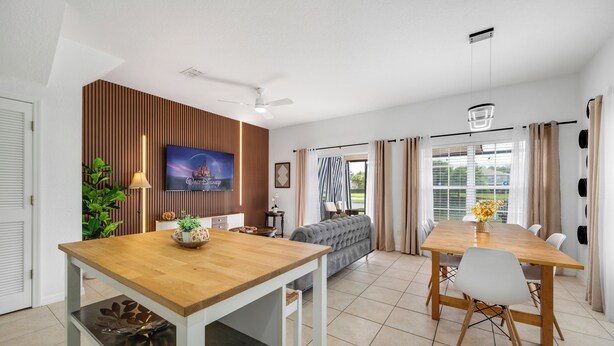 A modern living room with a wooden dining table, a sofa, a TV, and a large window with curtains. - THEMED 4 BR Coral Cay GEM w/Lake view-10 min to Disney-Free BBQ-Free resort (Kissimmee)