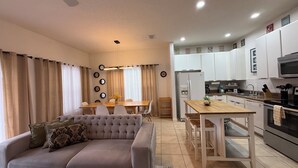 A modern living room with a sofa, dining table, and kitchen area with white cabinets and appliances. - THEMED 4 BR Coral Cay GEM w/Lake view-10 min to Disney-Free BBQ-Free resort (Kissimmee)