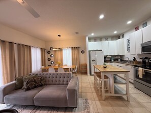 A modern living room with a sofa, dining table, and kitchen area with white cabinets and appliances. - THEMED 4 BR Coral Cay GEM w/Lake view-10 min to Disney-Free BBQ-Free resort (Kissimmee)