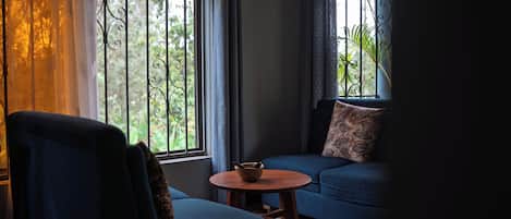 Una habitación con un sofá azul, una silla, una mesita redonda y una ventana con cortinas.