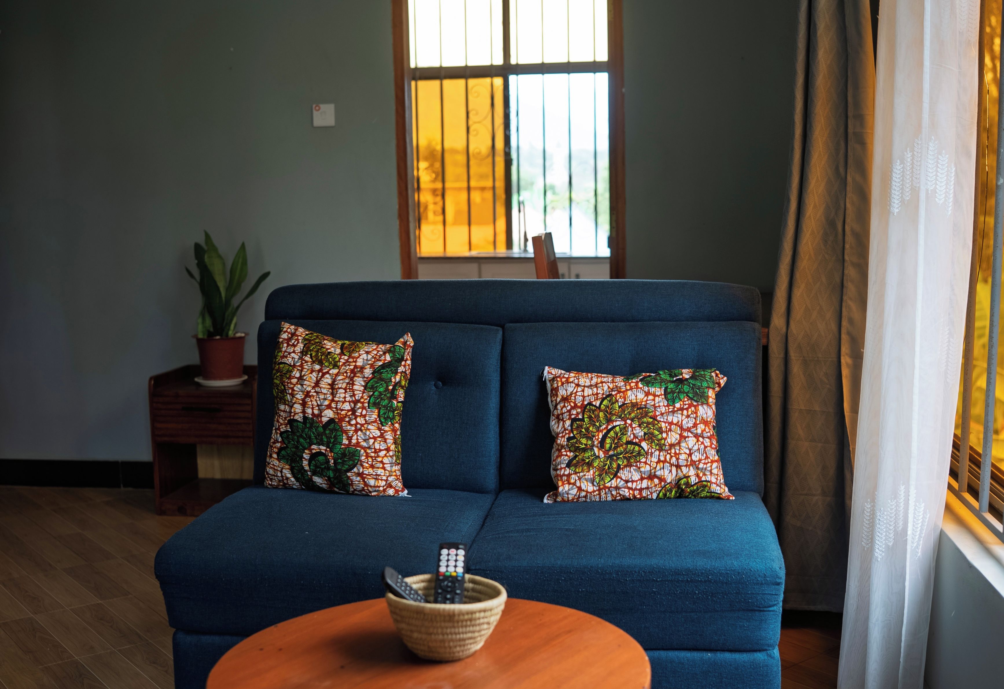 Ein blaues Sofa mit gemusterten Kissen, ein kleiner Holztisch mit einer Topfpflanze und ein gewebter Korb mit Fernbedienungen auf einem Holztisch.