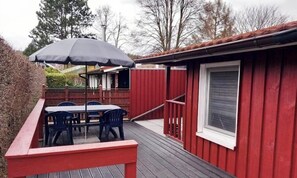 Eine rote Holzdielenveranda mit Tisch und Stühlen, ein überdachter Bereich mit Sonnenschirm und ein Fenster an einem Gebäude.