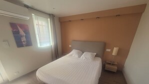A neatly made bed in a room with a wooden floor, a window with blinds, and a wall-mounted light fixture. - A 58 m² one-bedroom ground-floor apartment, detached and adjacent to a Provençal villa (Jonquières)