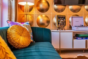 Ein blaustürkises Sofa mit einem gemusterten Kissen, eine dekorative Lampe, ein Plattenspieler und ein Bücherregal mit Büchern und einer Schallplatte.