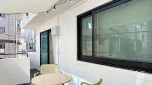 Un balcon avec une table et des chaises, une grande fenêtre et un mur en briques blanches.