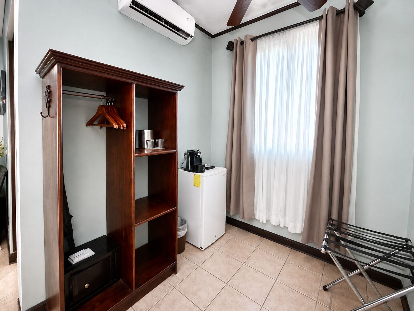 A hotel room with a wardrobe, a small kitchenette, a drying rack, and a window with curtains.