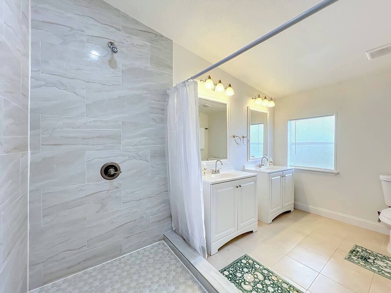 A bathroom with a shower, two sinks, a toilet, and a window.