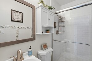 A bathroom with a white sink, a toilet, a shower with a glass door, a framed quote on the wall, and a shelf with plants. - 3-bedroom house in wonderful Winter Park with Big Fenced-In Back Yard! (Winter Park)