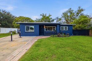 Una casa de un piso con fachada azul, una puerta principal y un pequeño jardín delantero con césped y plantas.