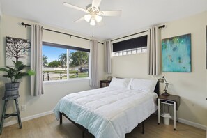 Ein Schlafzimmer mit einem Bett, einem Deckenventilator, einem Fenster mit Vorhängen, einem Nachttisch, einer Lampe und einer Topfpflanze.