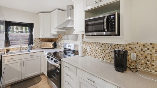 A compact kitchen with white cabinets, a built-in microwave, a stove, and a coffee maker. - 3-bedroom house in wonderful Winter Park with Big Fenced-In Back Yard! (Winter Park)
