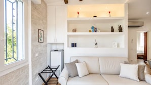 A modern living room with a white sofa, a wall-mounted shelf with decorative items, and a window with a view of greenery. - Casa nel Bosco – Families & Groups, Jacuzzi (Godiasco Salice Terme)