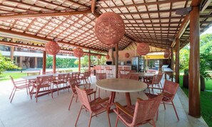 An outdoor patio area with round tables and wicker chairs, a brick fireplace, and a covered ceiling structure. - Ap w/ 2 Air-conditioned Suites Itacimirim Lnv0003 (Camaçari)