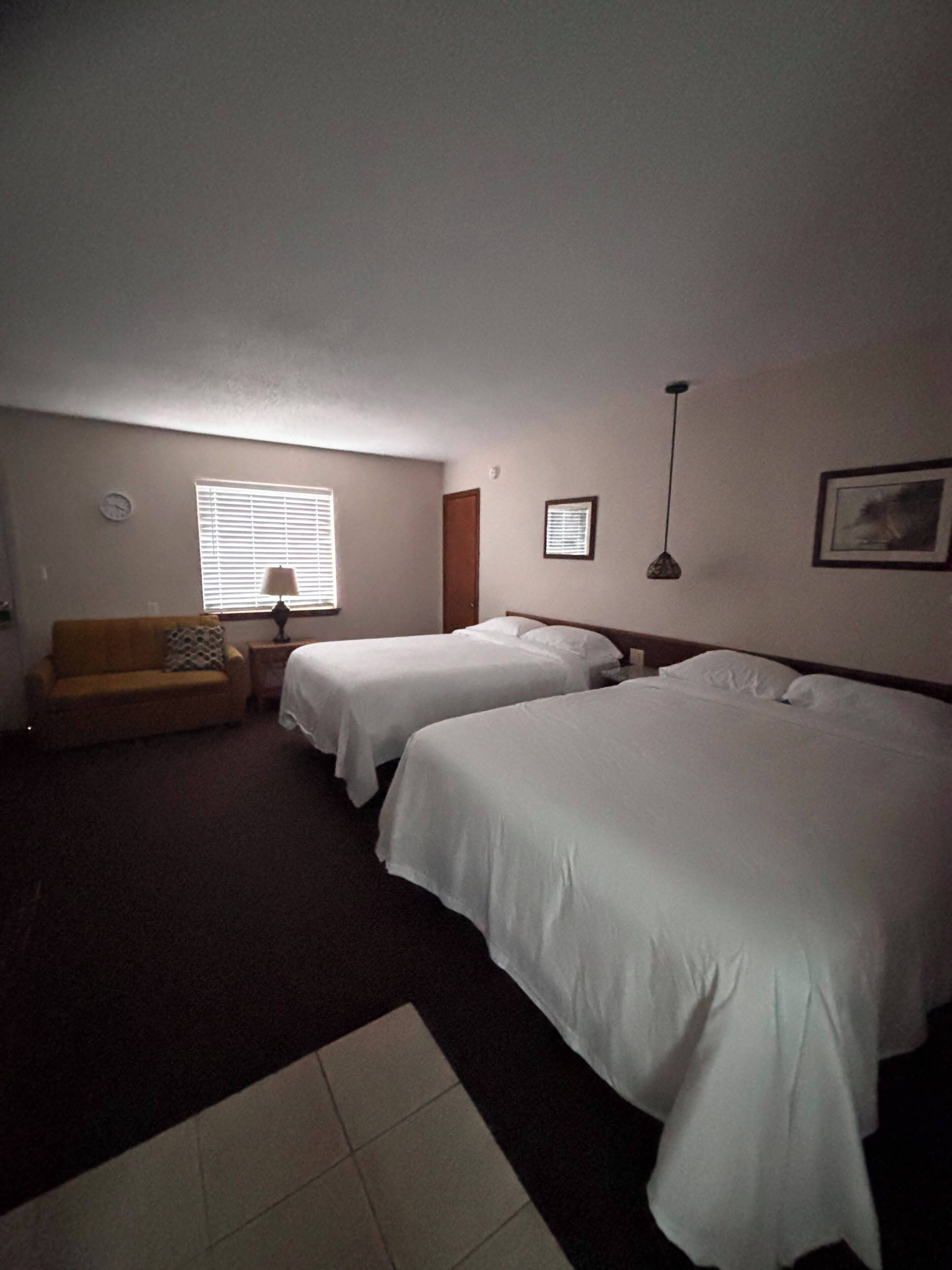 Habitación de hotel con dos camas individuales, un sofá, una mesita, una lámpara y una ventana con persianas.