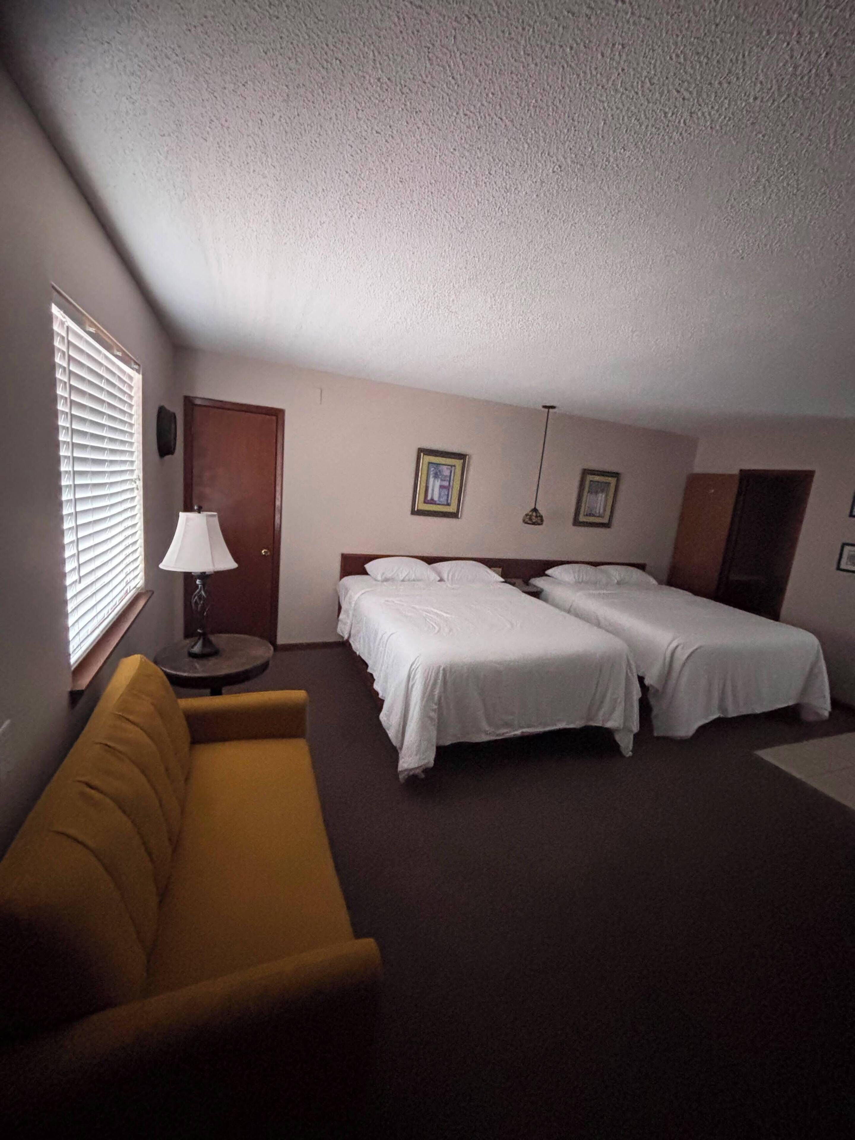 A hotel room with two beds, a sofa, a lamp, and a window with blinds.